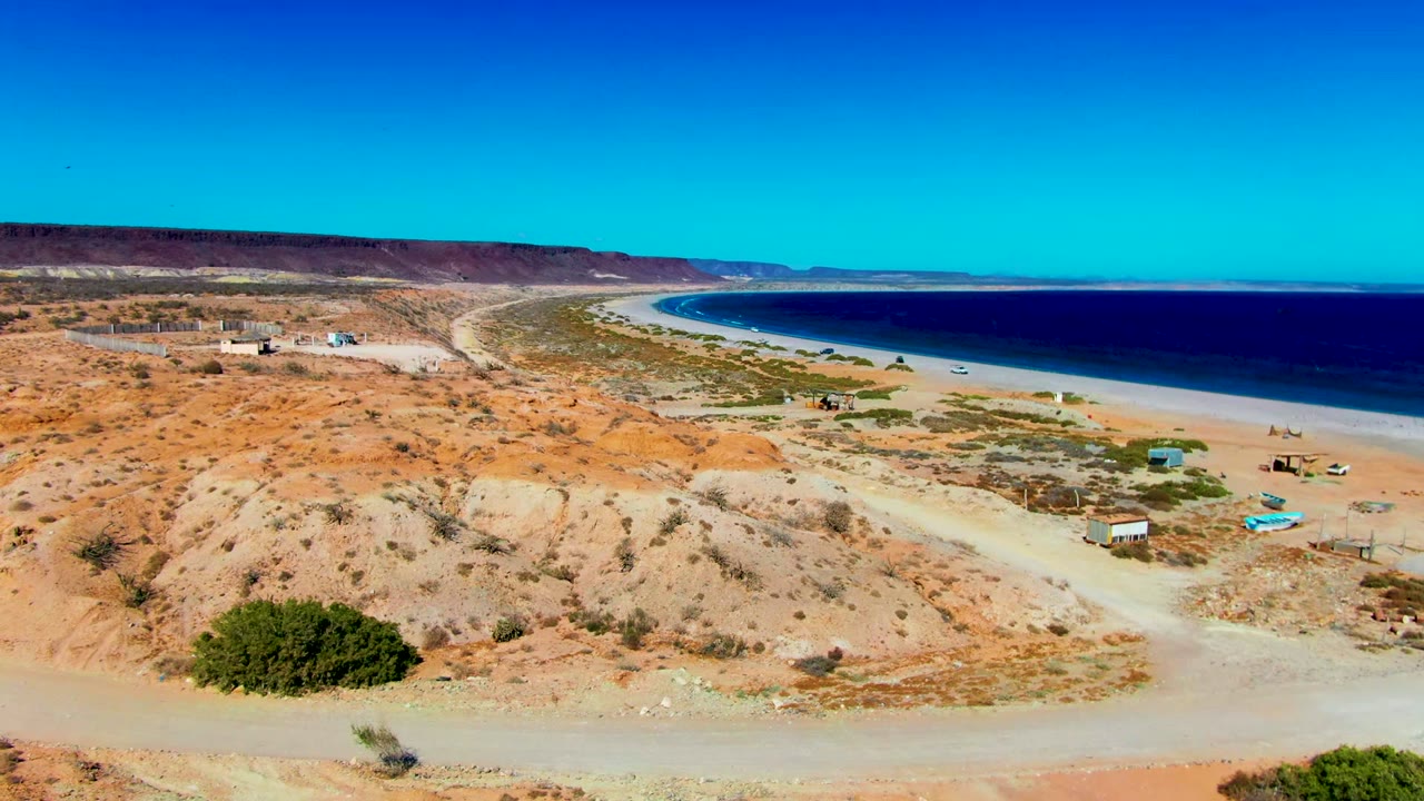 Aerial view over San Juanico, Baja California Sur, with orange desert scrub, a dirt road, small beach structures, curved shoreline, blue ocean, and no visible people.
