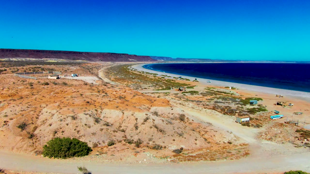 Wide aerial coastline with dirt roads, huts, vehicles, and ocean.