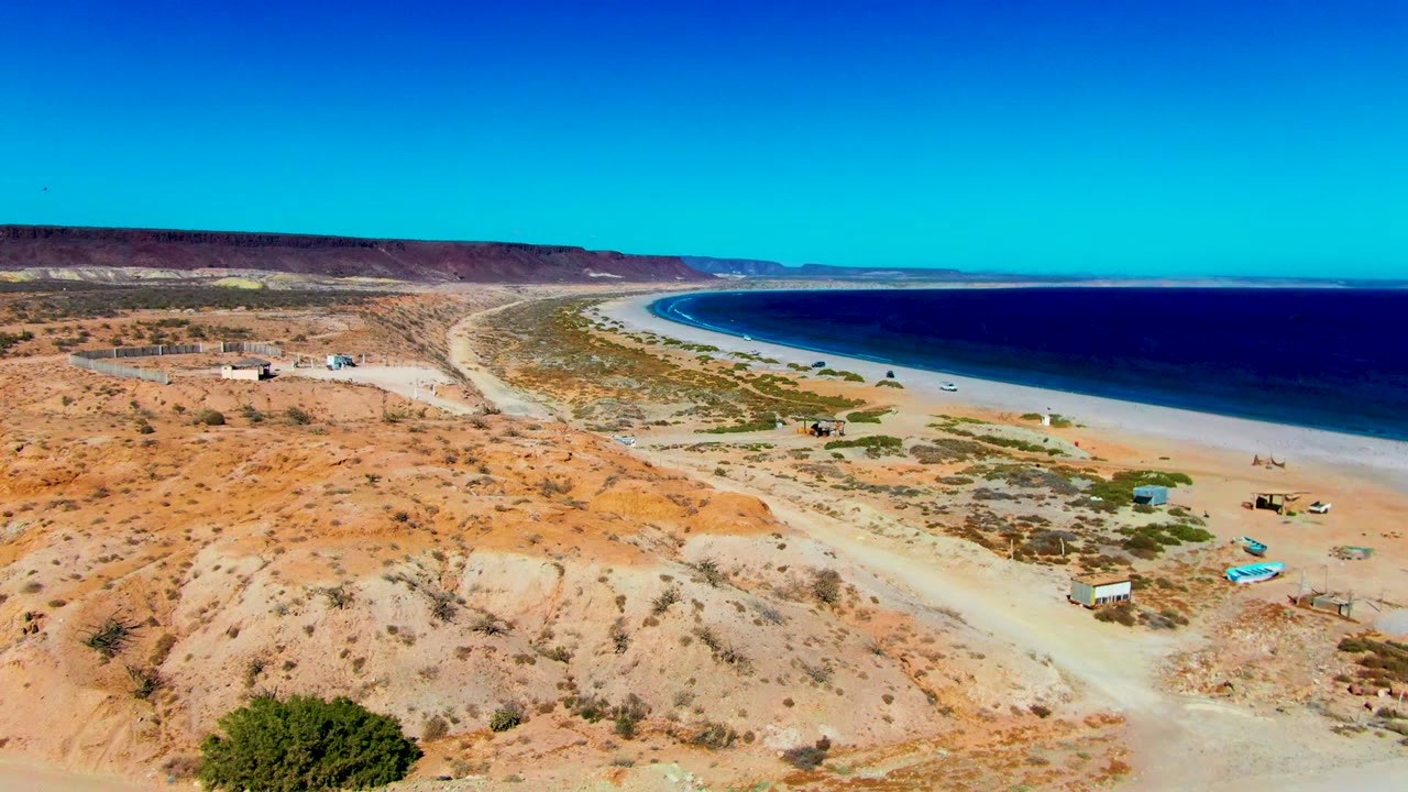 Aerial frame over San Juanico with a pale road crossing the desert foreground, vehicles near beach structures, a long sandy shoreline, blue ocean, and no visible people.