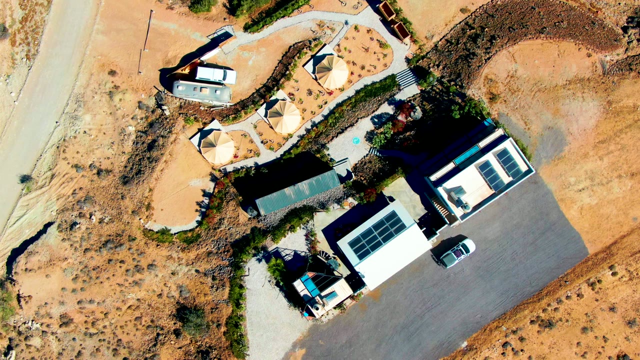 Static overhead view in San Juanico, Baja California Sur showing modern buildings, gray paved surfaces, a pale road, shade tents, sandy terrain, sparse greenery, a parked vehicle, and no visible people.
