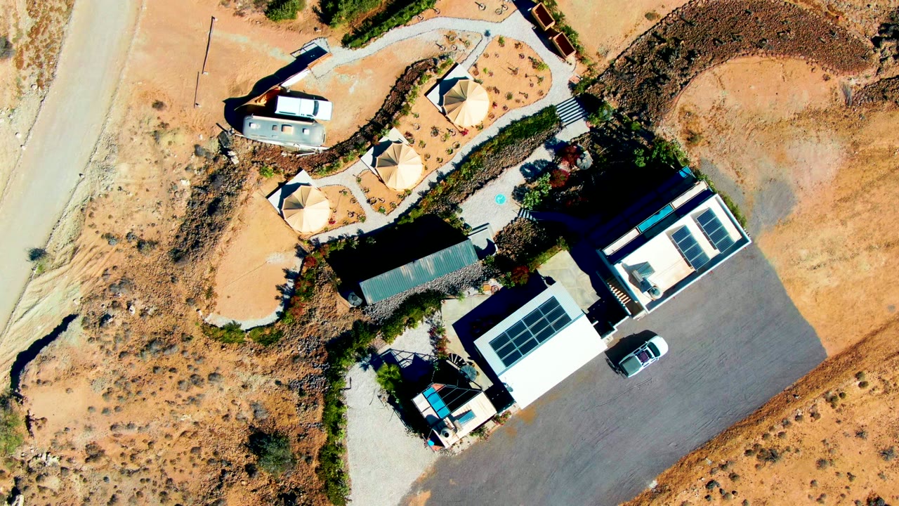 Top-down aerial frame over a San Juanico desert property, showing solar panels on modern buildings, a paved lot, a road, circular shade structures, a trailer, sandy terrain, and no visible people.