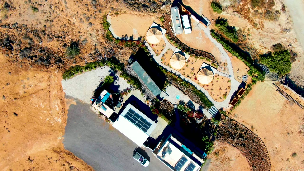 Aerial frame from San Juanico with the paved lot sweeping across the lower area, solar panels on two buildings, a trailer near the sandy road, circular shade structures, arid ground, and no visible people.