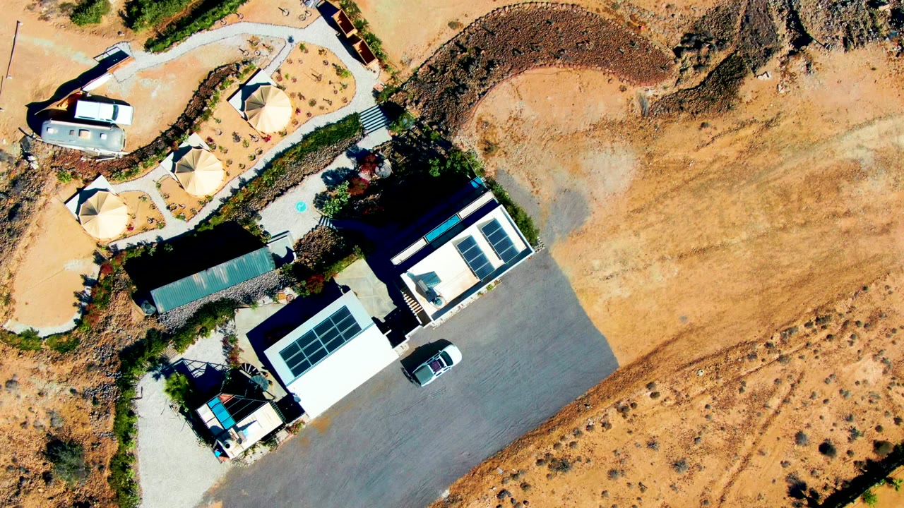 Overhead frame of a San Juanico desert building complex with roof solar panels, circular shade domes, paved parking, a parked vehicle, sandy terrain, and no visible people.