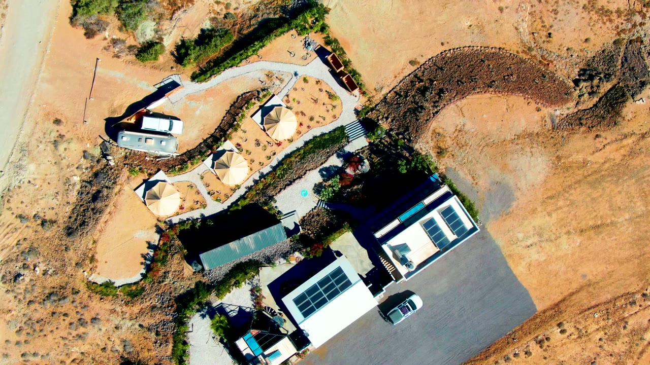 Top-down frame in San Juanico with the complex beside a pale road, solar-panel roofs, circular domes, dark pavement, scrub patches, and no visible people.