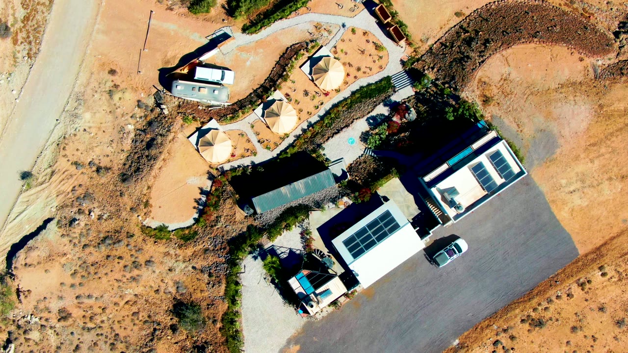 At 0:00, a top-down aerial frame shows a desert property in San Juanico, Baja California Sur with a white car near light-roofed buildings, circular shade structures, sandy roads, and no visible people.