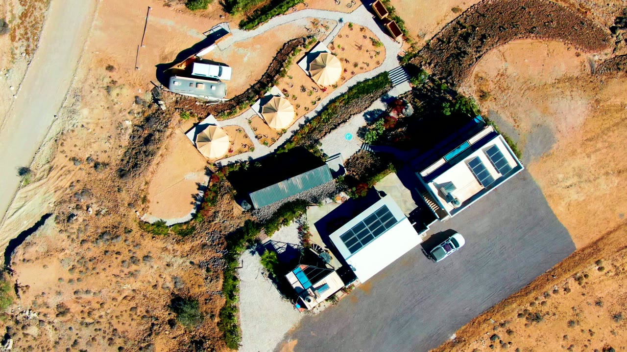 Overhead desert property view with buildings, circular structures, road, and a white car in the lot.