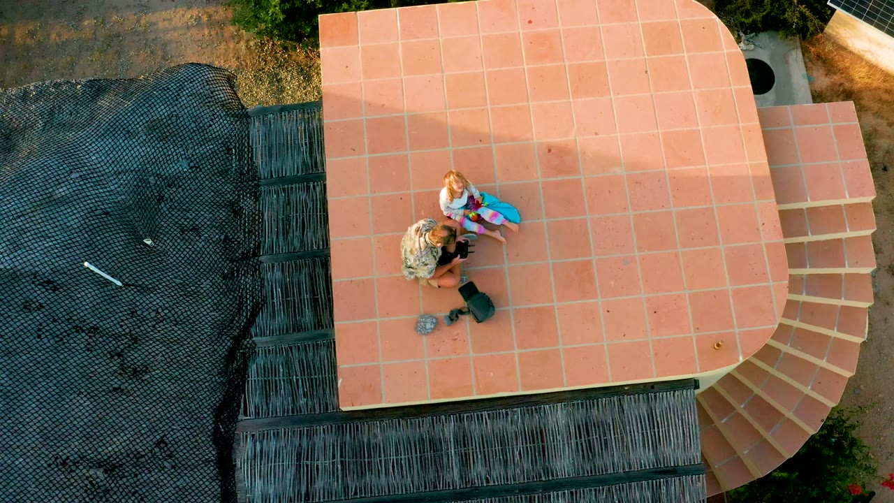 Top-down aerial frame in Cabo Pulmo, Baja California Sur, showing two people on a terracotta tiled rooftop beside a darker thatched roof edge.