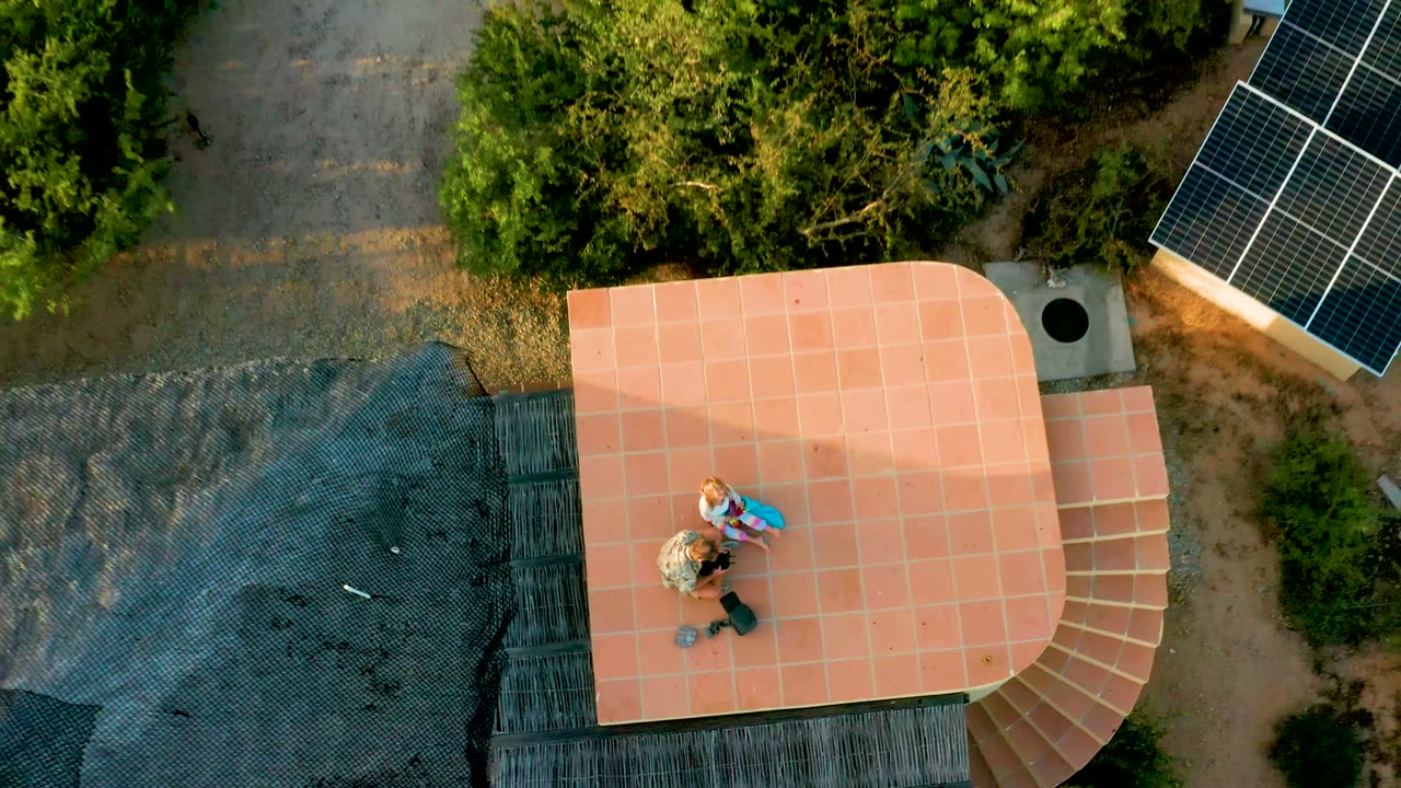 Wider top-down view in Cabo Pulmo showing two people on the tiled rooftop, a curved stair section, surrounding trees, dirt ground, and solar panels at the upper right.