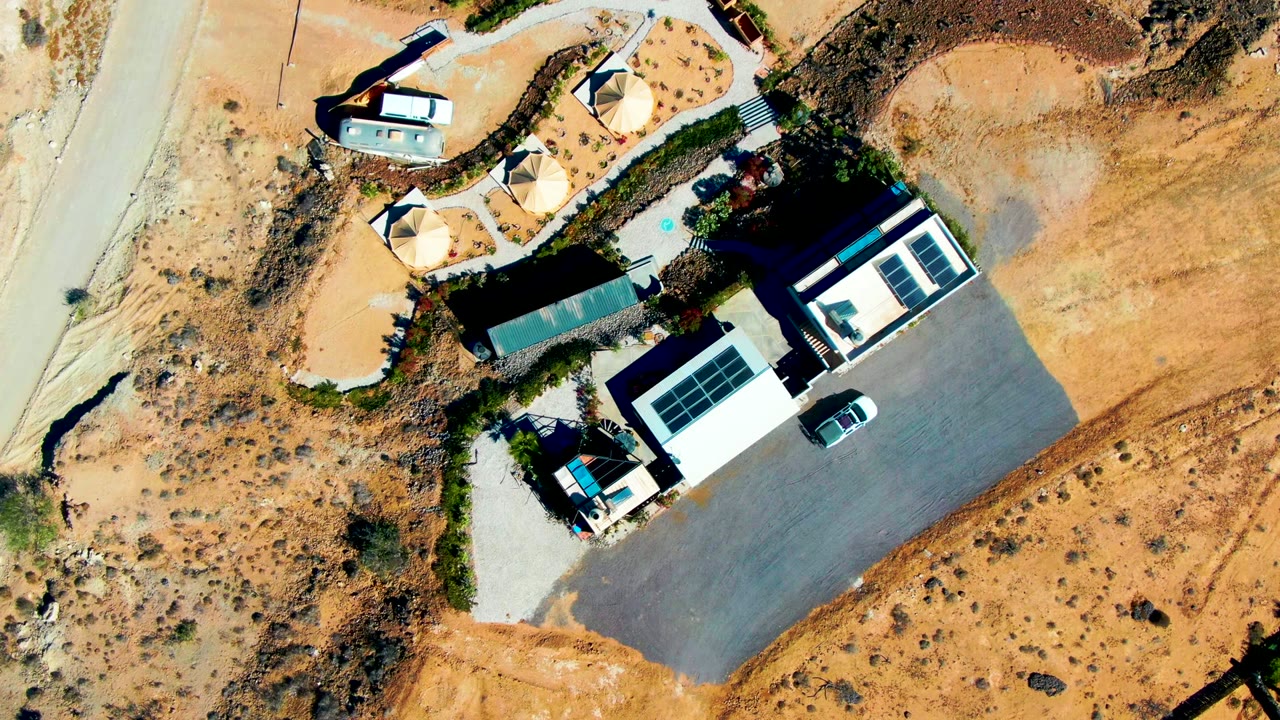 Overhead frame in San Juanico, Baja California Sur showing white-roofed buildings with solar panels, a parked car, sandy desert ground, a road edge, and no visible people.