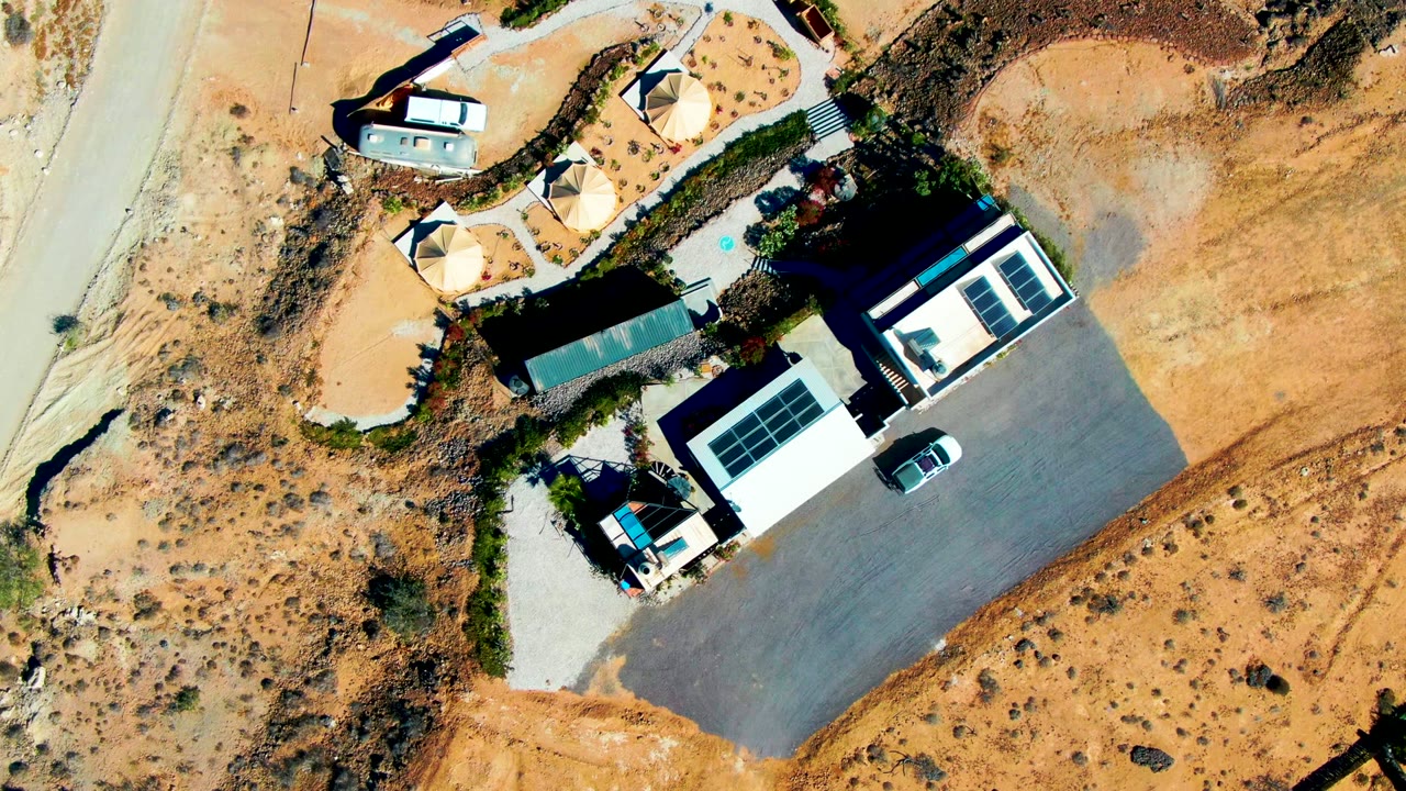 Aerial view of buildings, solar panels, a car, and dry terrain beside a road.