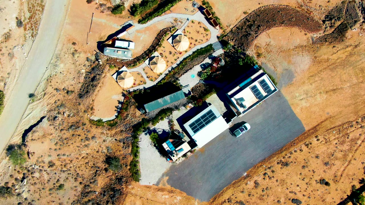 High overhead view of a San Juanico desert property with modern roofed structures, solar panels, a curved dirt road, sandy scrubland, parked vehicles, and no visible people.
