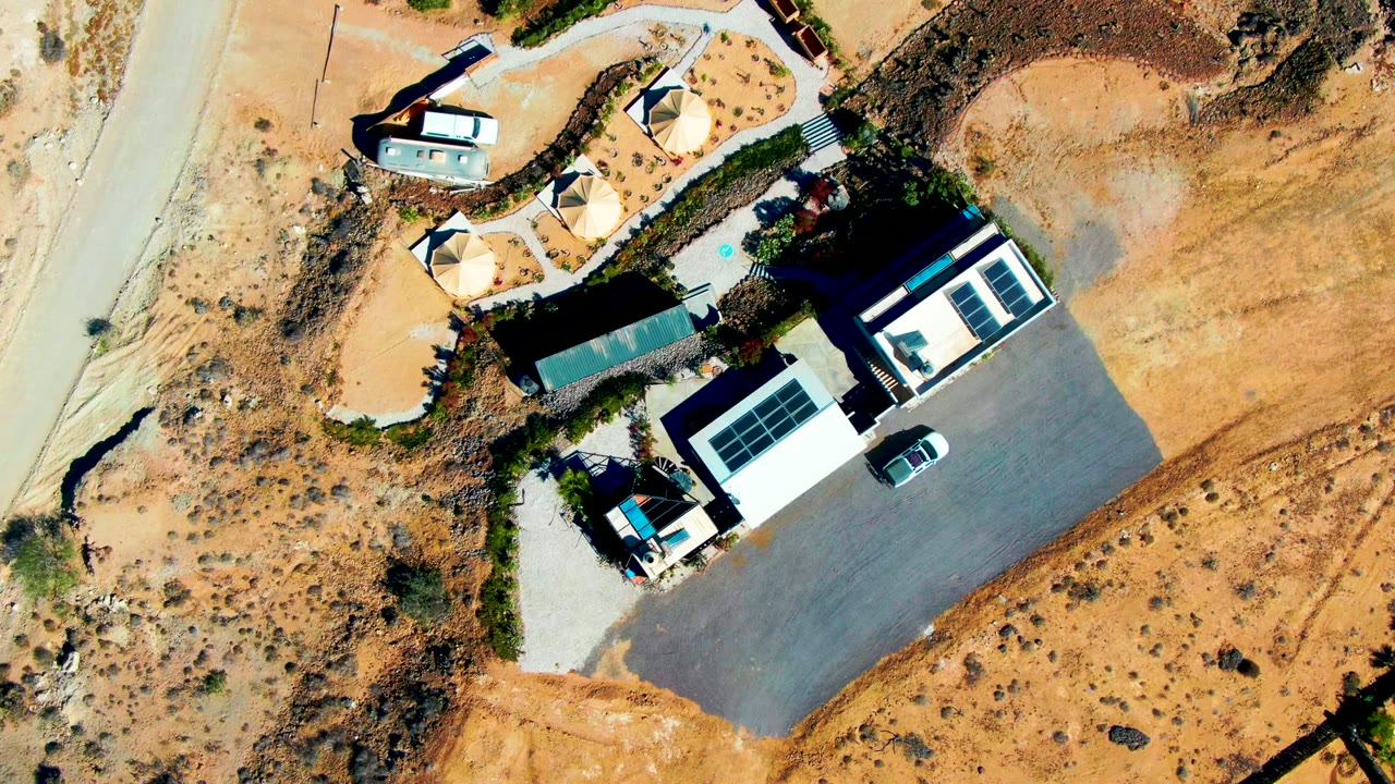 Top-down San Juanico frame with the road along the left side, shade canopies near the upper building, solar panels on lower roofs, open sandy ground, vehicles, and no visible people.