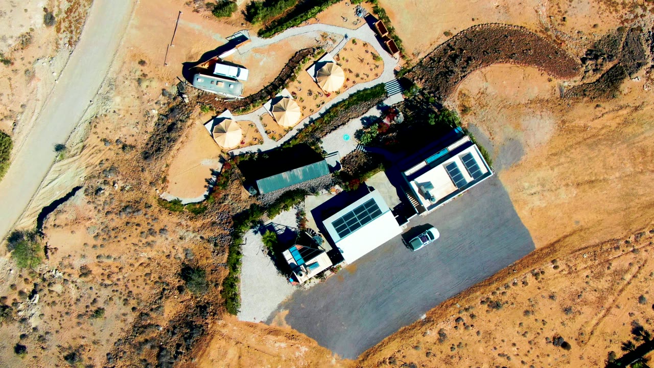 Overhead drone frame of a San Juanico desert property with white-roofed buildings, shade structures, a paved parking lot, sandy scrubland, a pale road, and no visible people.