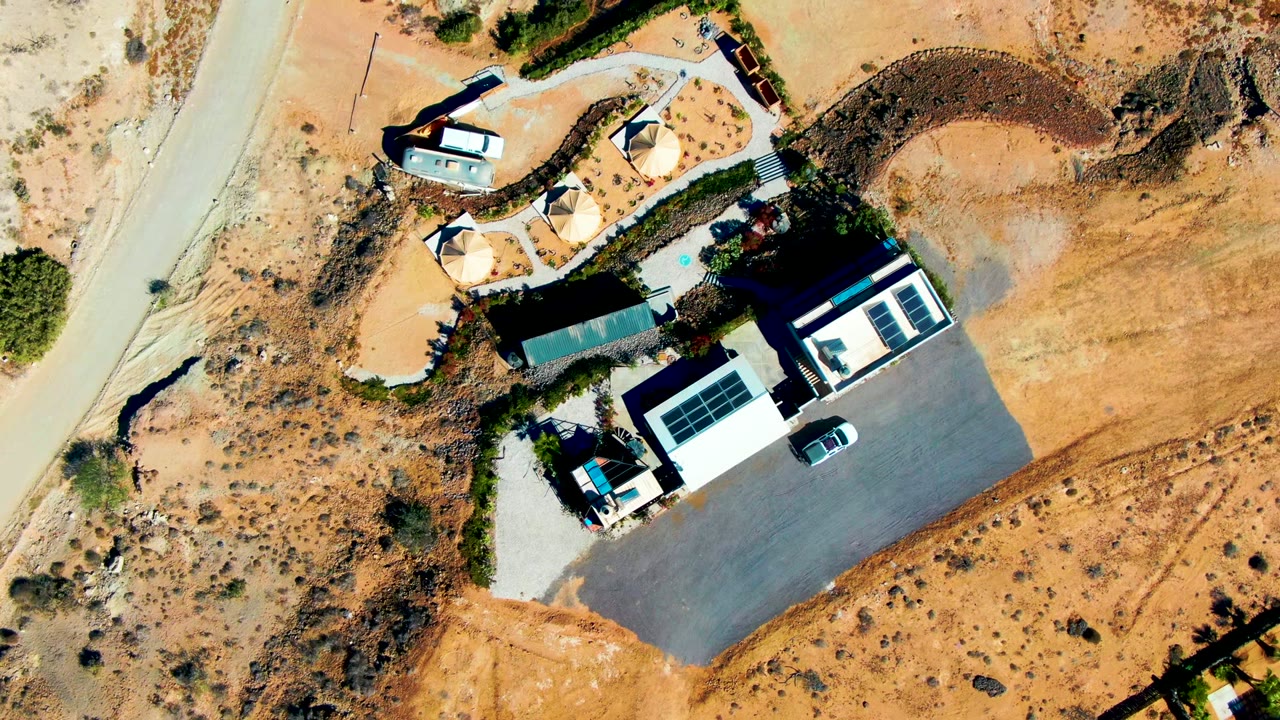 Top-down aerial frame in San Juanico, Baja California Sur, showing a pale road beside sandy terrain, a small building compound, shade tents, parked vehicles, and no visible people.