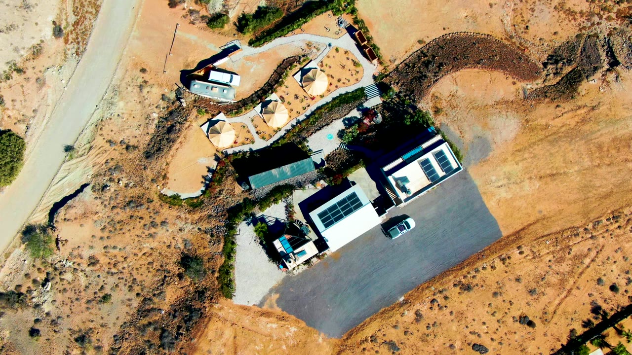 Aerial frame from San Juanico, Baja California Sur, showing a small desert property, light-roofed structures, a dark paved area, tan open ground, scattered bushes, and no visible people.
