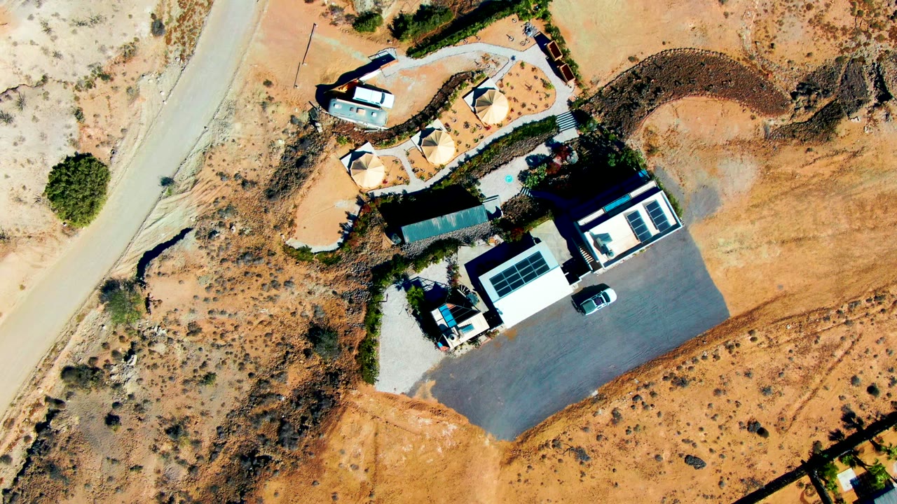 Overhead view in San Juanico, Baja California Sur, Mexico showing a desert compound beside a curved road, small huts, a solar-panel roof, a parked vehicle, and a tiny person near the upper path.
