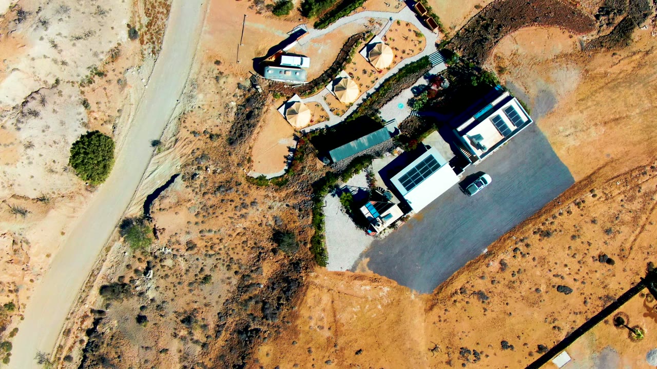 High overhead frame of a San Juanico desert compound with a paved parking area, solar-panel roof, shade structures, sandy ground, sparse vegetation, and no visible people.