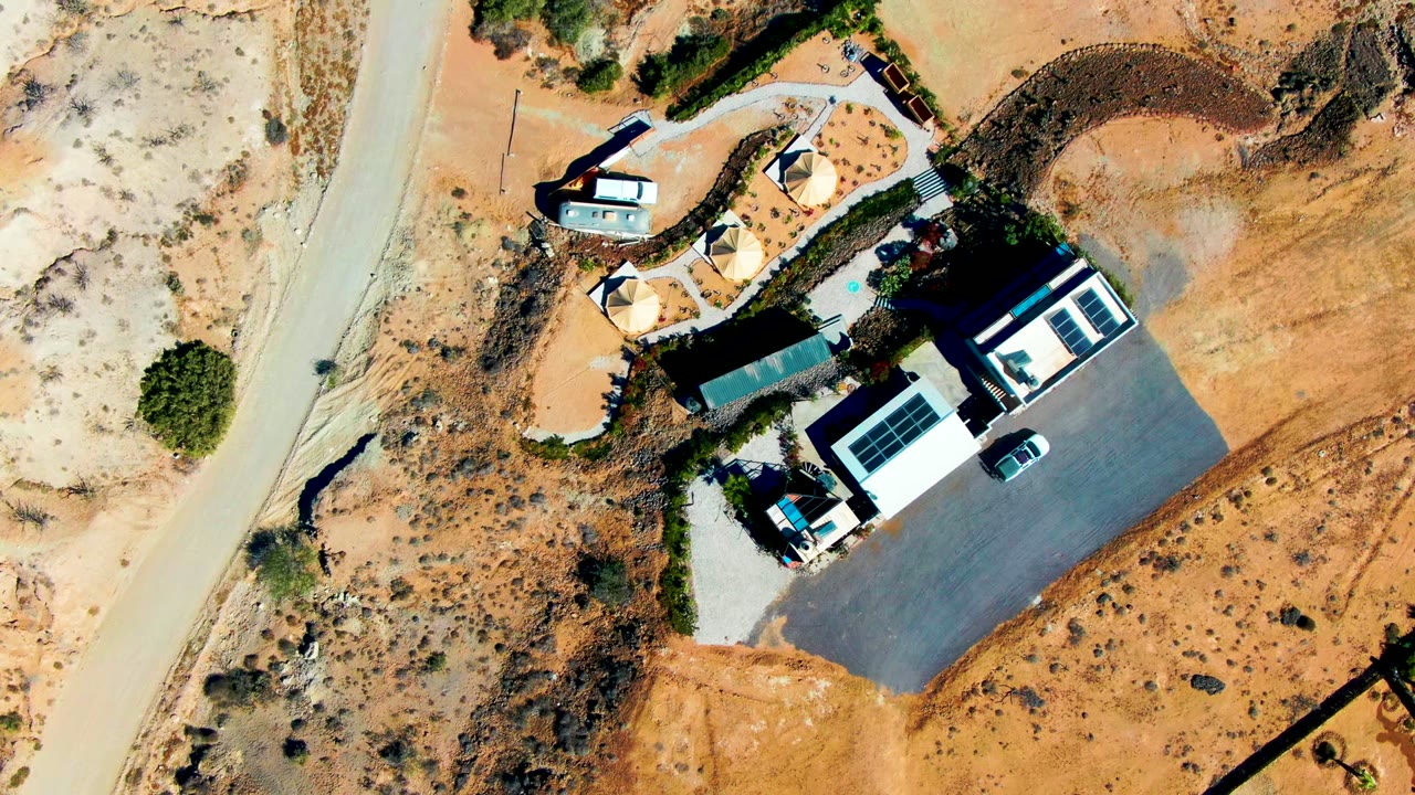 Near-vertical frame over San Juanico desert ground with the main roofed building, adjacent shade structures, a parked vehicle, a paved lot, and no visible people.