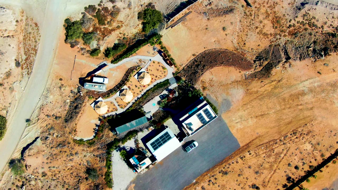 Top-down view in San Juanico, Baja California Sur showing a white building, dark parking area, curved road, small vehicles, shade structures, dry sand, sparse shrubs, and no visible people.