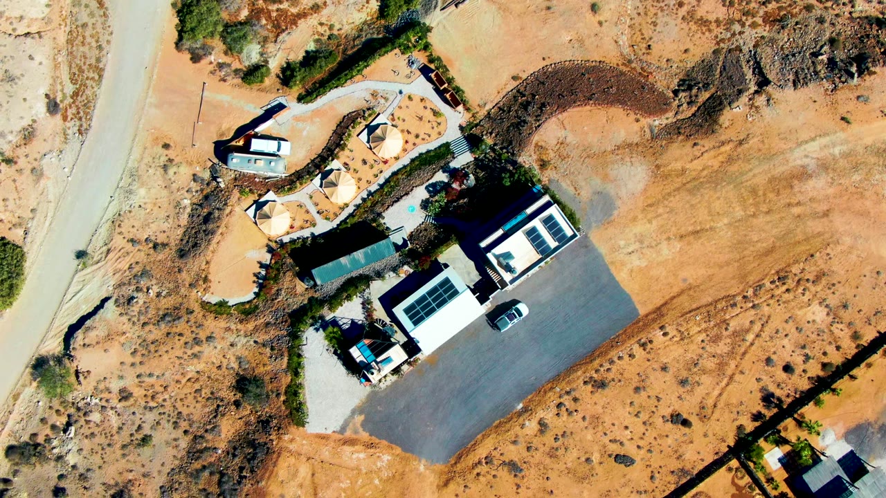 Aerial frame over San Juanico with a white desert building, dark paved parking, two small vehicles, tent-like shade forms, a road along the left edge, dry brush, and no visible people.