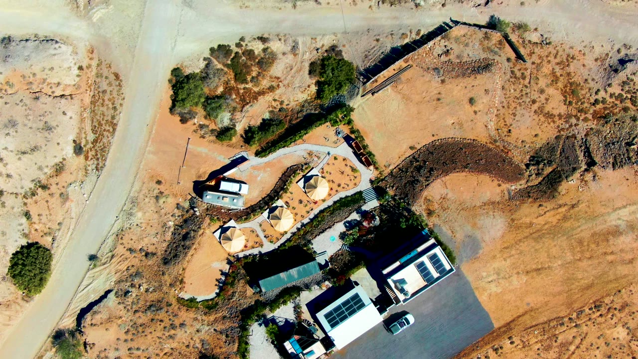 Top-down aerial frame in San Juanico, Baja California Sur showing a pale dirt road, modern house, driveway, shade structures, sparse bushes, and no visible people.