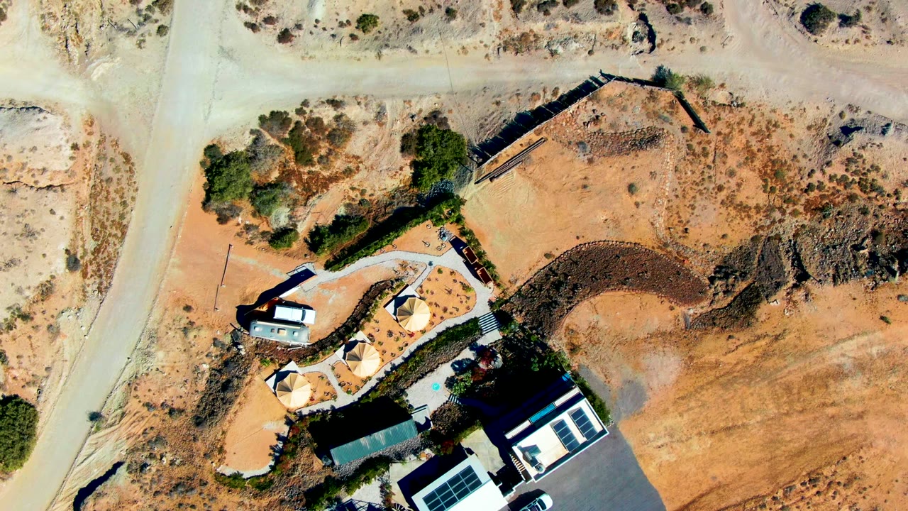 San Juanico aerial frame with the dirt road along the left side, the house and paved driveway below, and a broad dry lot with scattered shrubs to the right, no visible people.