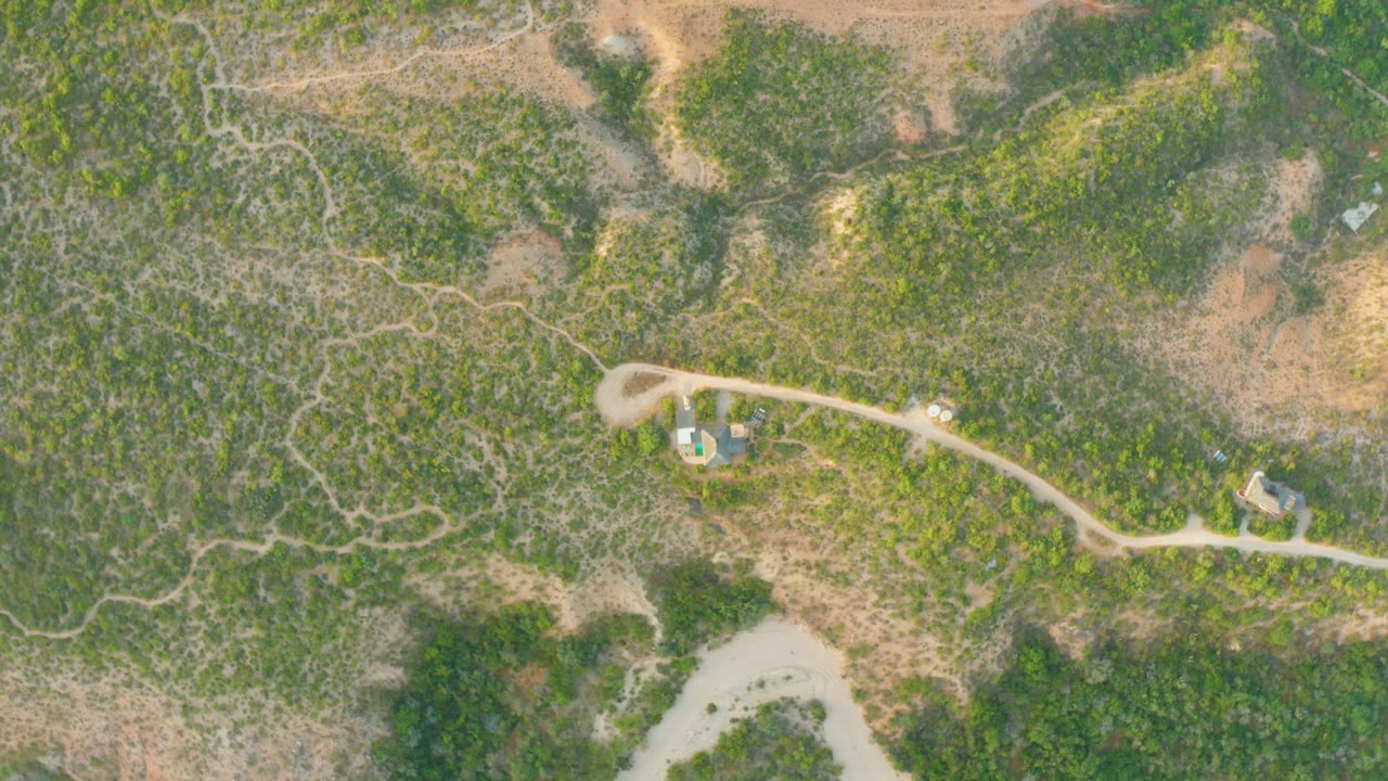 Overhead frame from Cabo Pulmo, Baja California Sur showing a pale dirt road through scrubland, a small blue-roofed structure near the road, another small building to the right, and no visible people.