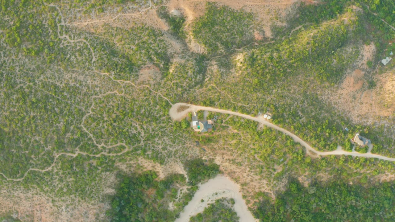 Overhead Cabo Pulmo frame with a light-colored vehicle on the pale road, scattered green brush, dry brown patches, a blue-roofed structure near the bend, and no visible people.