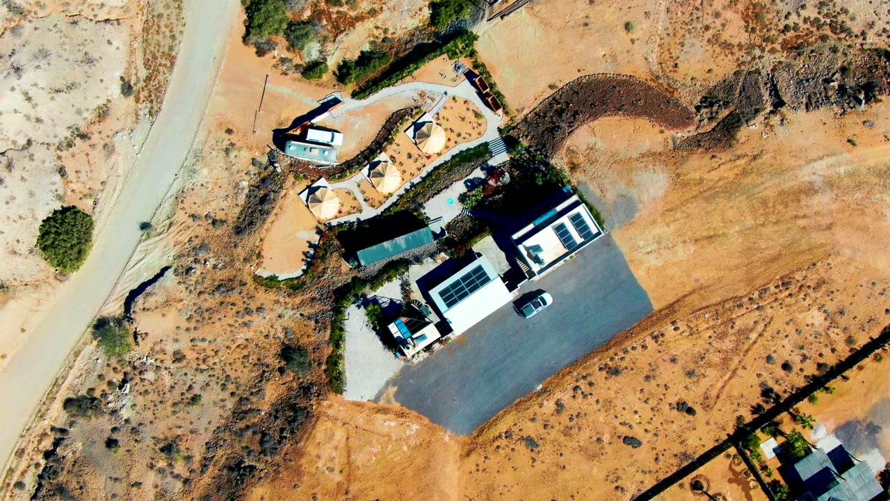 Aerial frame of a white-roofed home beside a road in dry desert terrain.