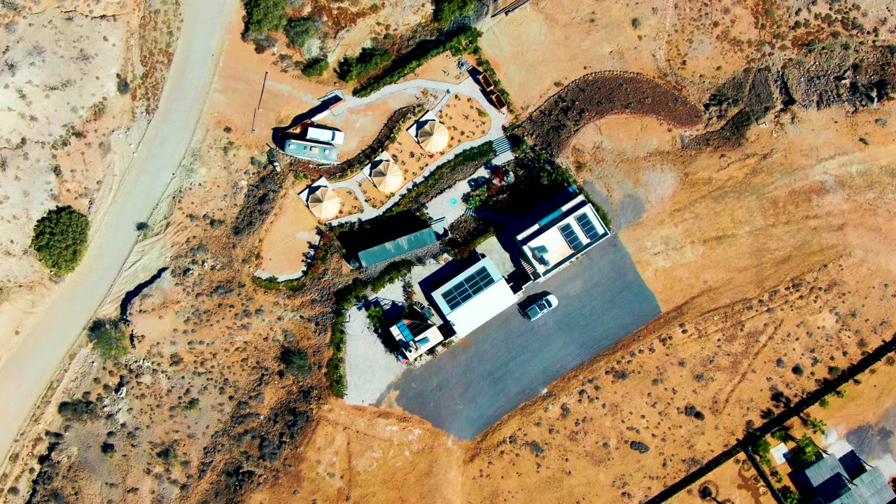 Top-down San Juanico frame showing the residential property, parked car on the driveway, pale road at left, curved landscaped area above the house, dry brush, and no visible people.