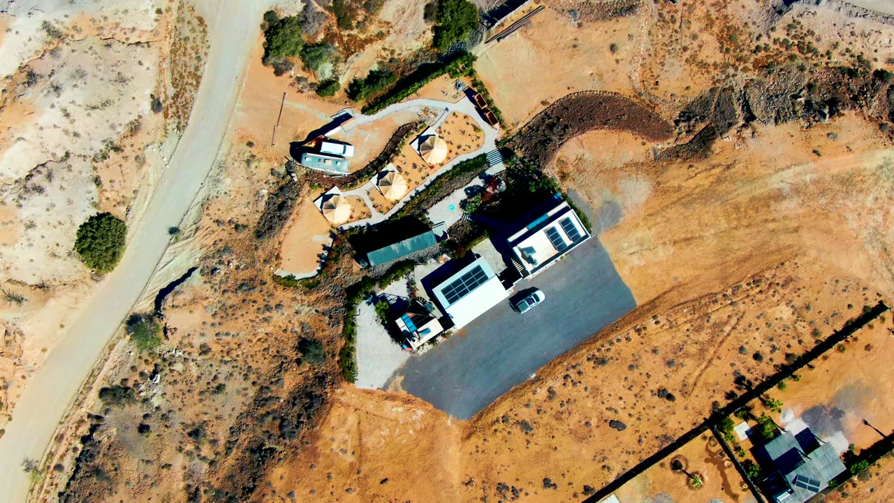 Opening overhead frame of a house compound in San Juanico, Baja California Sur, with a curved dirt road, pale desert ground, sparse vegetation, a driveway, a parked white vehicle, and no visible people.