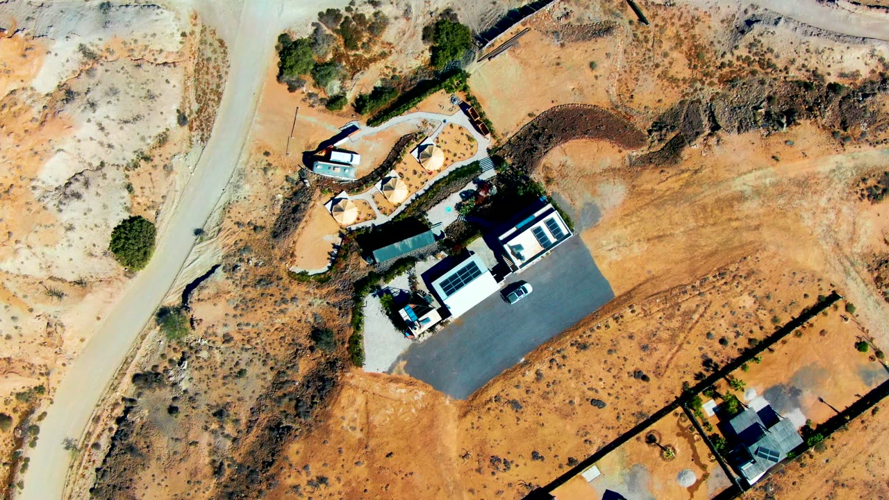Overhead drone frame in San Juanico, Baja California Sur showing a small car near the upper left road, dry scrubland, fenced lots, and houses with no visible people.