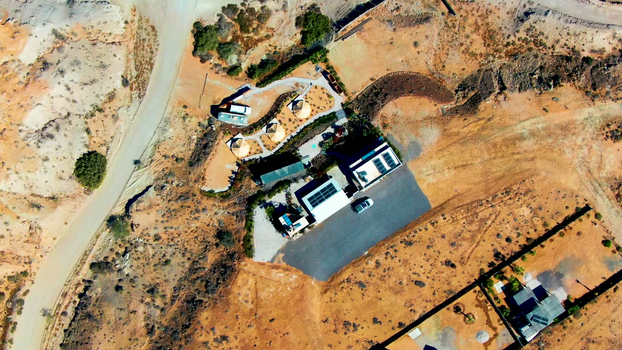 High overhead view in San Juanico, Baja California Sur with the car slightly farther along the left road, open tan ground, walled homes, and no visible people.