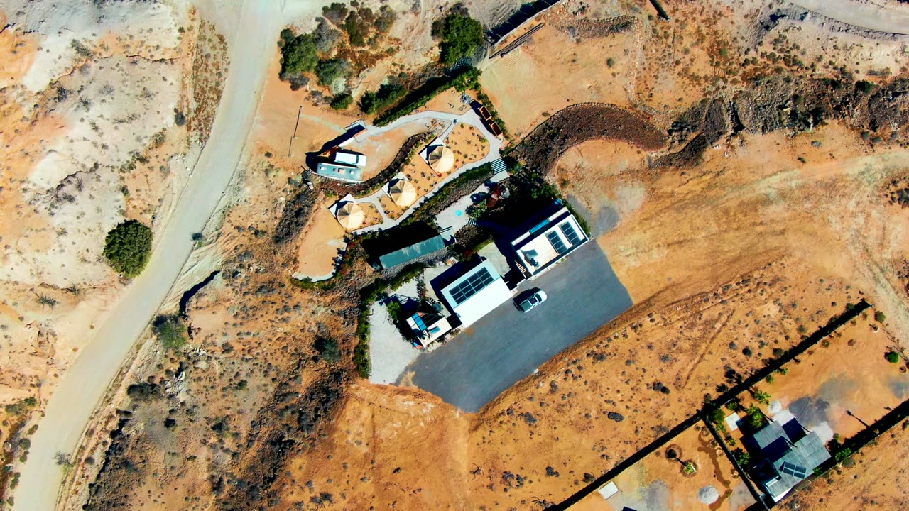 Top-down frame over San Juanico, Baja California Sur showing the car on the left-side road, nearby houses, bare brown lots, scattered shrubs, and no visible people.