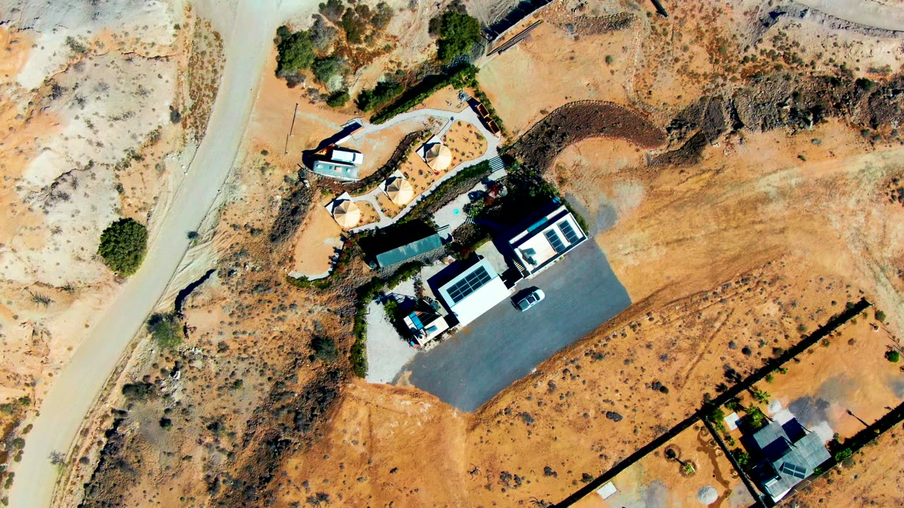 Drone view of a dry road, car, houses, and open desert ground.