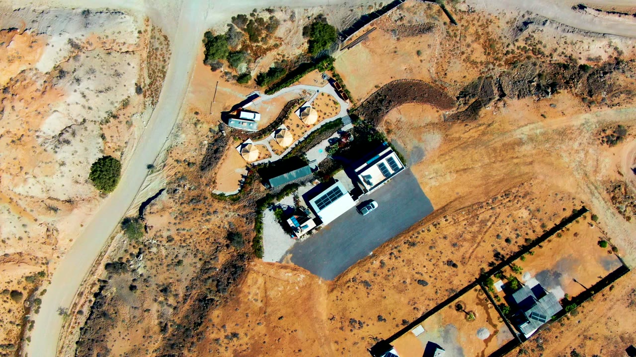 Overhead frame of a small residential compound in San Juanico with dark roof shadows, a curved access road, fenced sandy yards, scattered scrub, and no visible people.