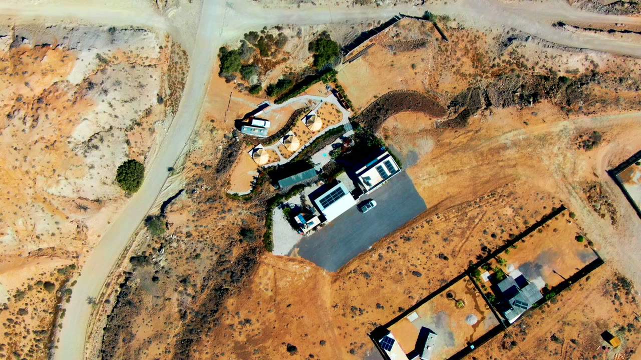 Overhead drone frame of a dry construction area in San Juanico, Baja California Sur, with pale dirt roads, walled lots, roofed structures, parked vehicles, scrub, and no visible people.