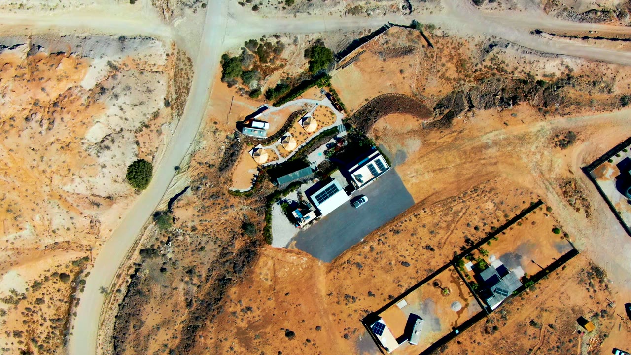 Top-down aerial frame over San Juanico, Baja California Sur showing sandy lots, dirt roads, small buildings, concrete pads, sparse scrub, and no visible people.