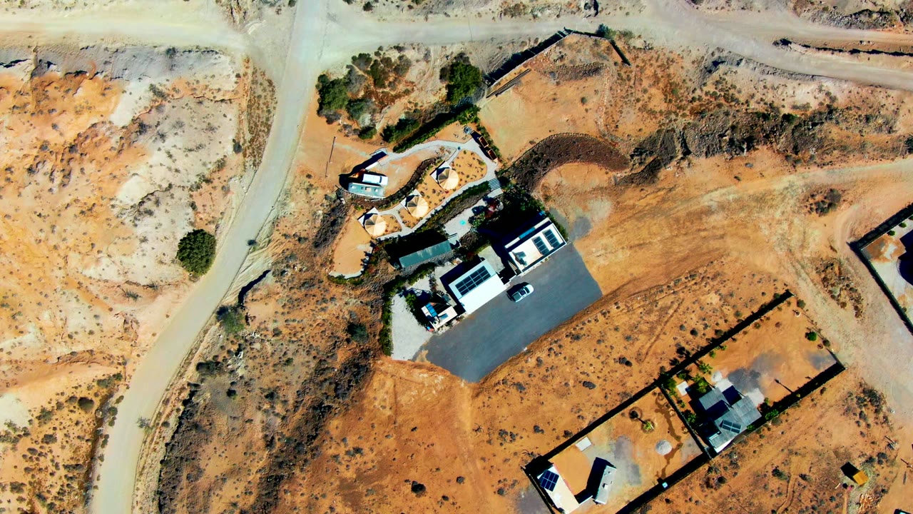 Overhead frame over San Juanico, Baja California Sur with unfinished lots, a dark rectangular surface, several roofed structures, pale road edges, and no visible people.