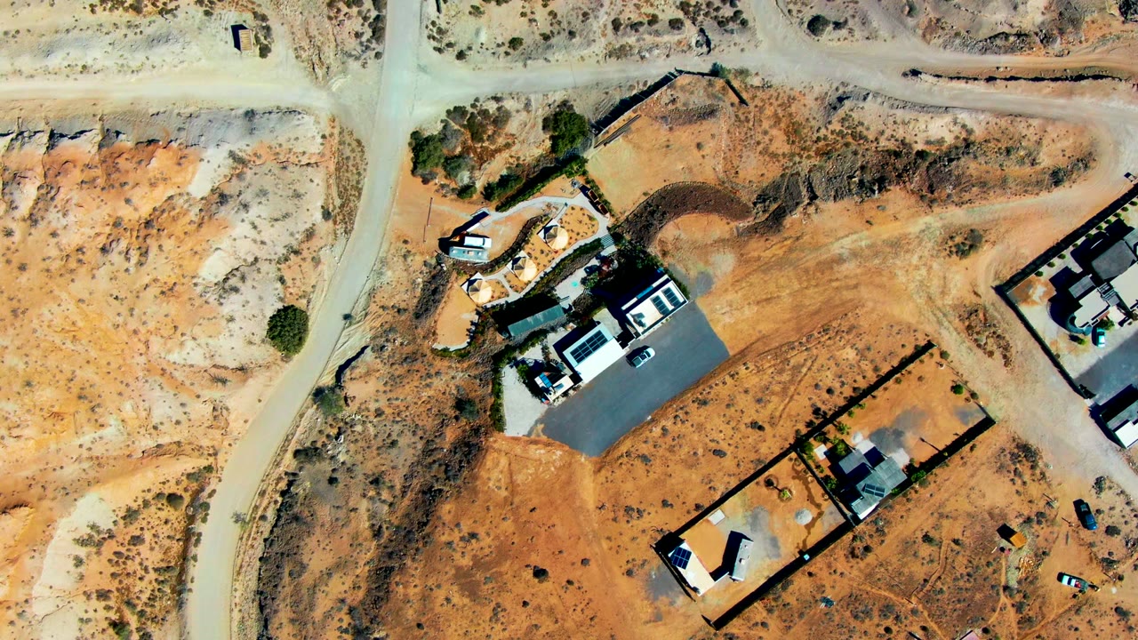 Aerial view of a dry road junction, fenced plots, buildings, and a vehicle on dusty ground.