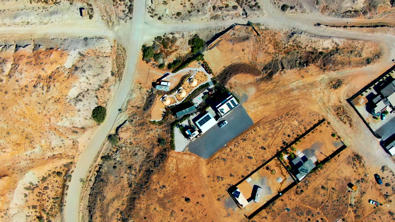 Aerial frame in San Juanico, Baja California Sur with dusty roads crossing dry terrain, multiple buildings, fenced rectangular lots, scattered shrubs, and small vehicles visible from above, with no visible people.