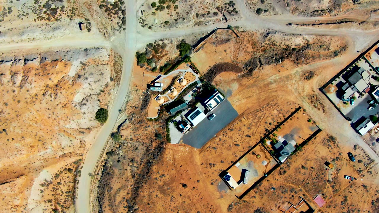 Overhead drone frame in San Juanico, Baja California Sur showing pale dirt roads, orange desert ground, walled lots, partially built houses, parked vehicles, and no visible people.