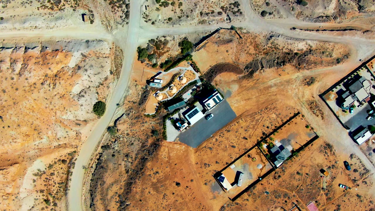 Top-down San Juanico frame showing tan dirt roads crossing dry orange ground, several fenced lots, partially built homes, parked vehicles, and no visible people.