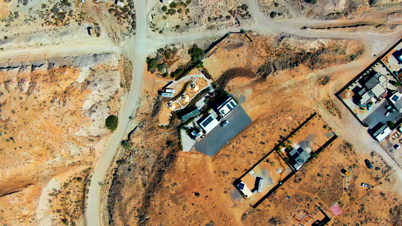 Top-down aerial frame over San Juanico, Baja California Sur showing dirt roads, walled houses, sparse desert scrub, parked vehicles, and no visible people at the opening moment.