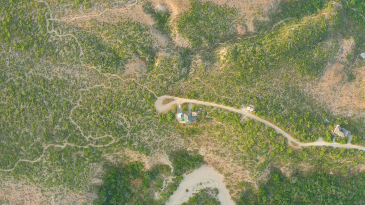 High aerial view in Cabo Pulmo, Baja California Sur, with a pale dirt road crossing scrubby hills, dry patches, small structures, and no visible people.