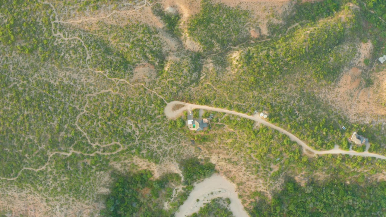 Aerial view over Cabo Pulmo hilly terrain, with the dirt road passing near small buildings, a light vehicle on the road, green vegetation, dry ground, and no visible people.