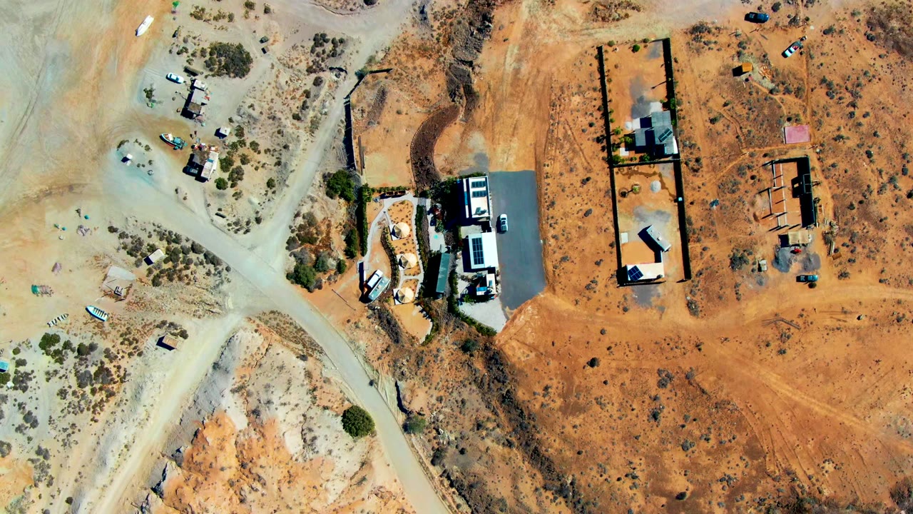 Overhead view of San Juanico desert lots, pale dirt roads, small buildings, sandy orange ground, sparse shrubs, and no visible people.