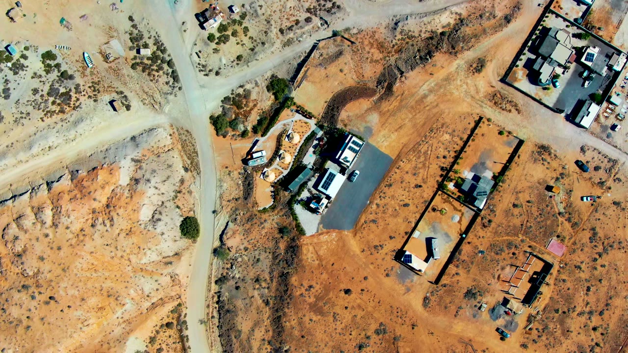 Aerial frame over San Juanico showing a pale road junction, orange earth, construction lots, dark roof shadows, scattered vehicles, and no visible people.