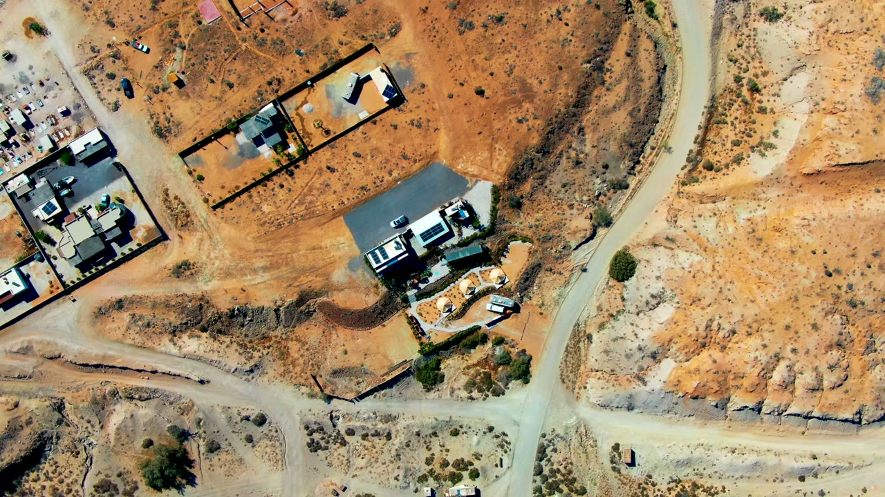Overhead aerial frame of San Juanico, Baja California Sur showing orange desert lots, pale dirt roads, walled homes, scattered shrubs, and no visible people.