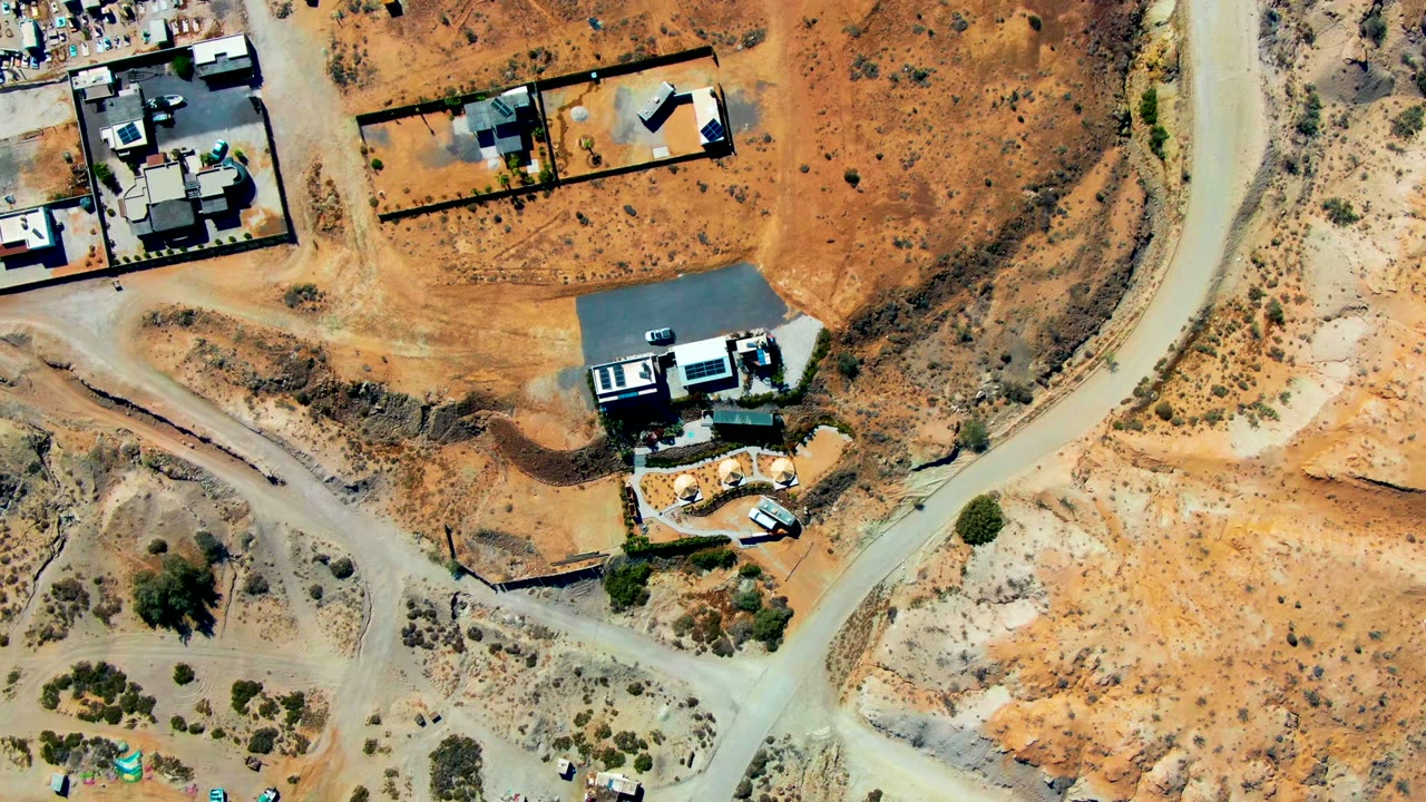 Top-down aerial view of a house beside a curving dirt road in desert terrain.